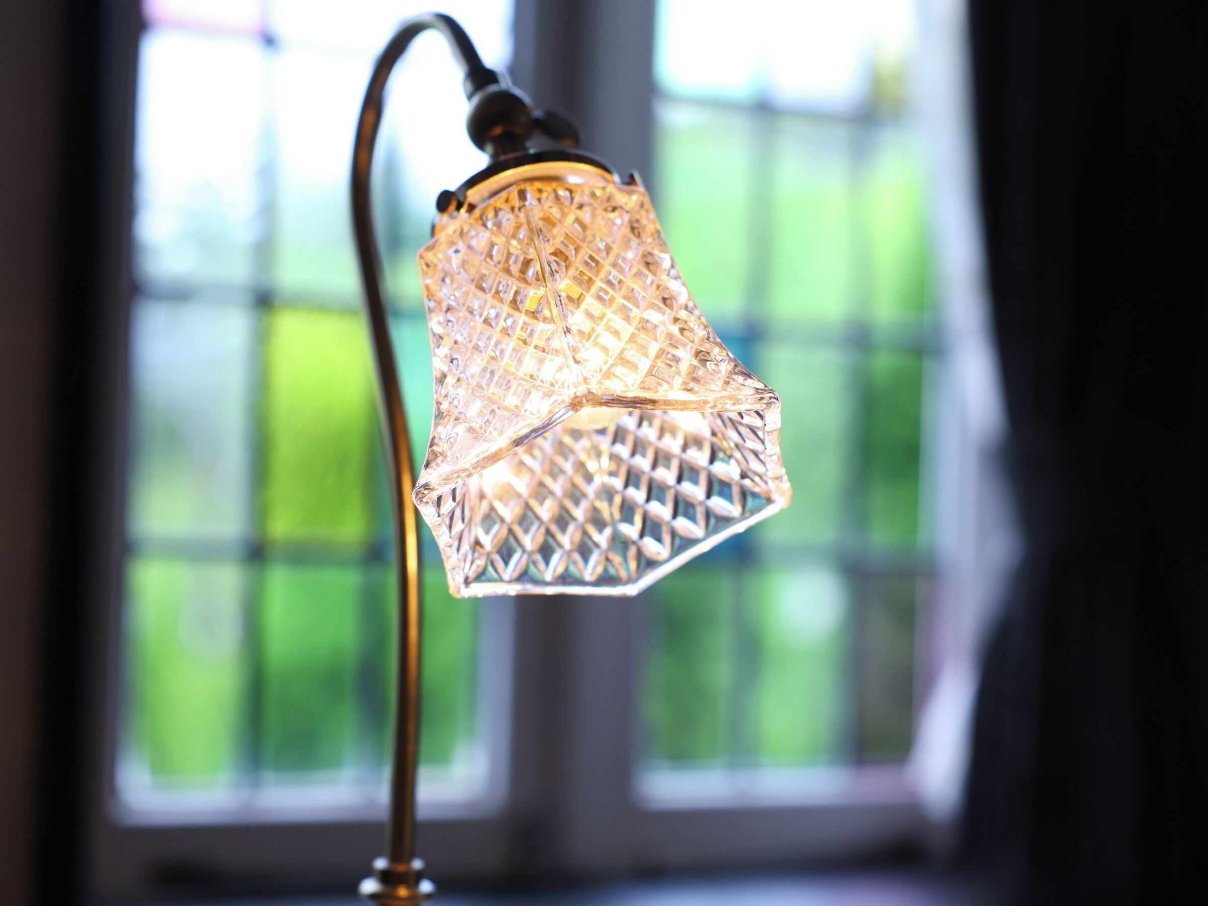 A close-up of a crystal lamp with a curved neck and a textured glass shade, positioned in front of a window with a blurred green outdoor view.