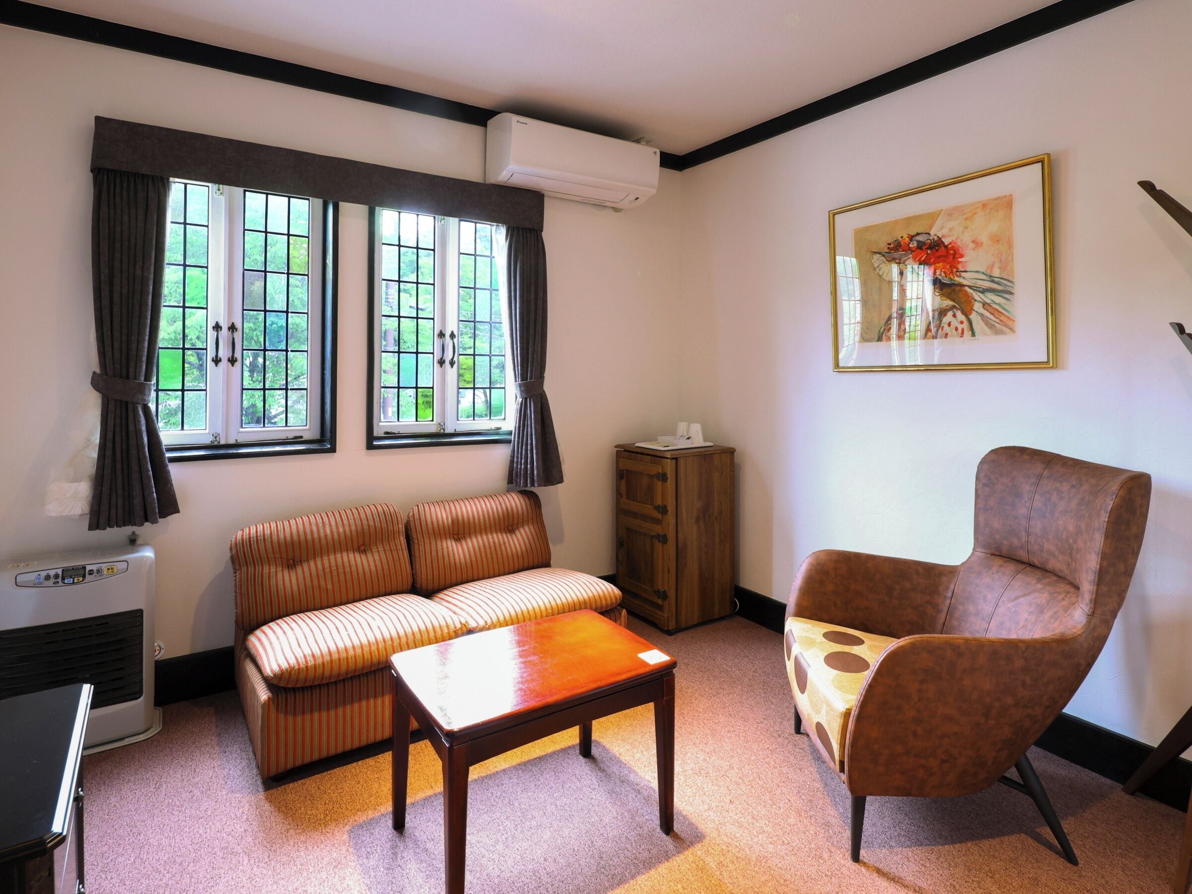 A living room with a sofa, armchair, and a small table. There are two windows with curtains, an air conditioning unit, and a framed picture on the wall.