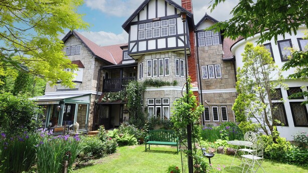 Una casa de estilo Tudor tradicional con fachada de piedra, múltiples ventanas y un jardín bien cuidado con un banco y plantas en macetas.