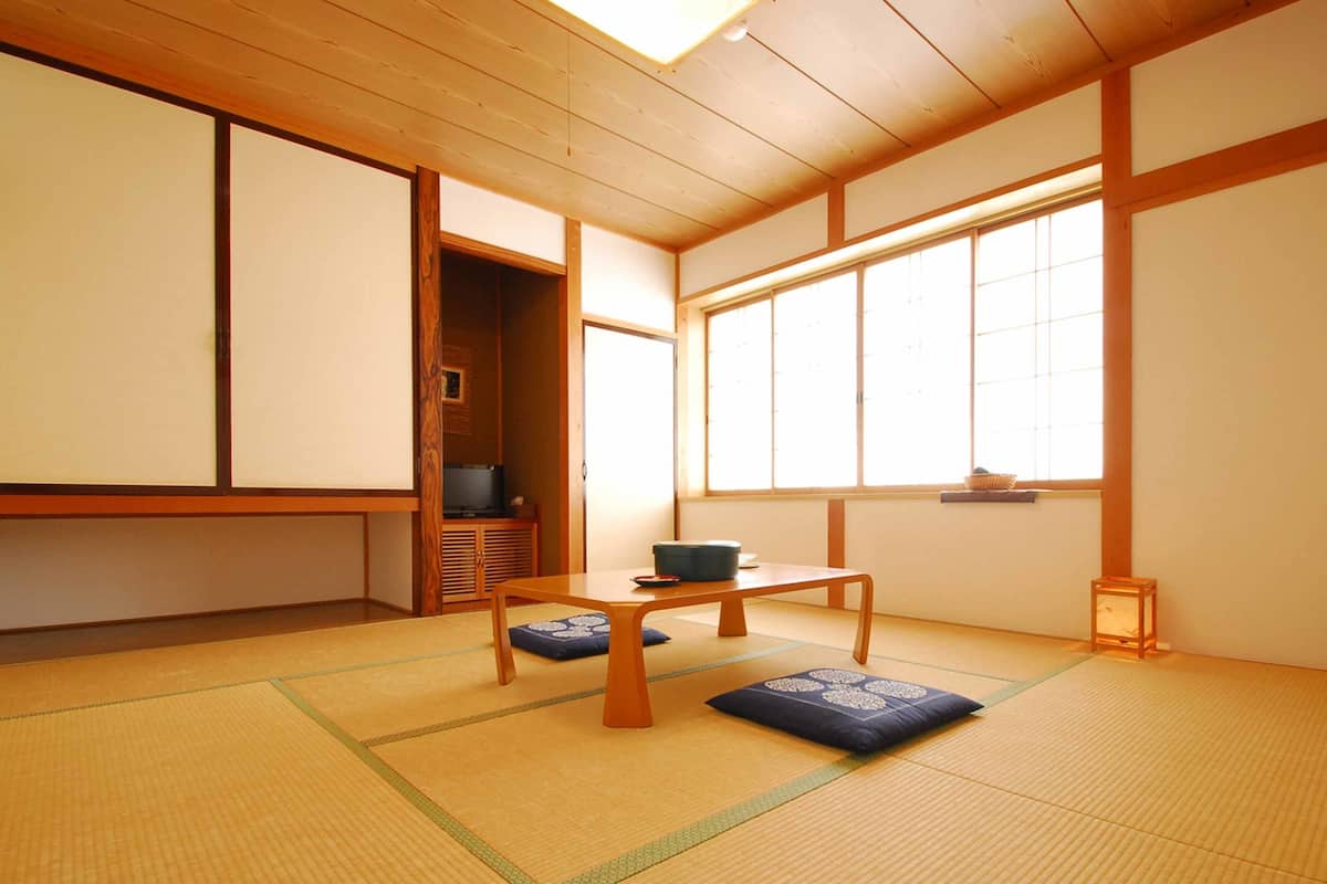 Een traditionele Japanse kamer met tatami vloerbedekking, een lage tafel met kussens, een televisie in een houten kast en grote ramen.