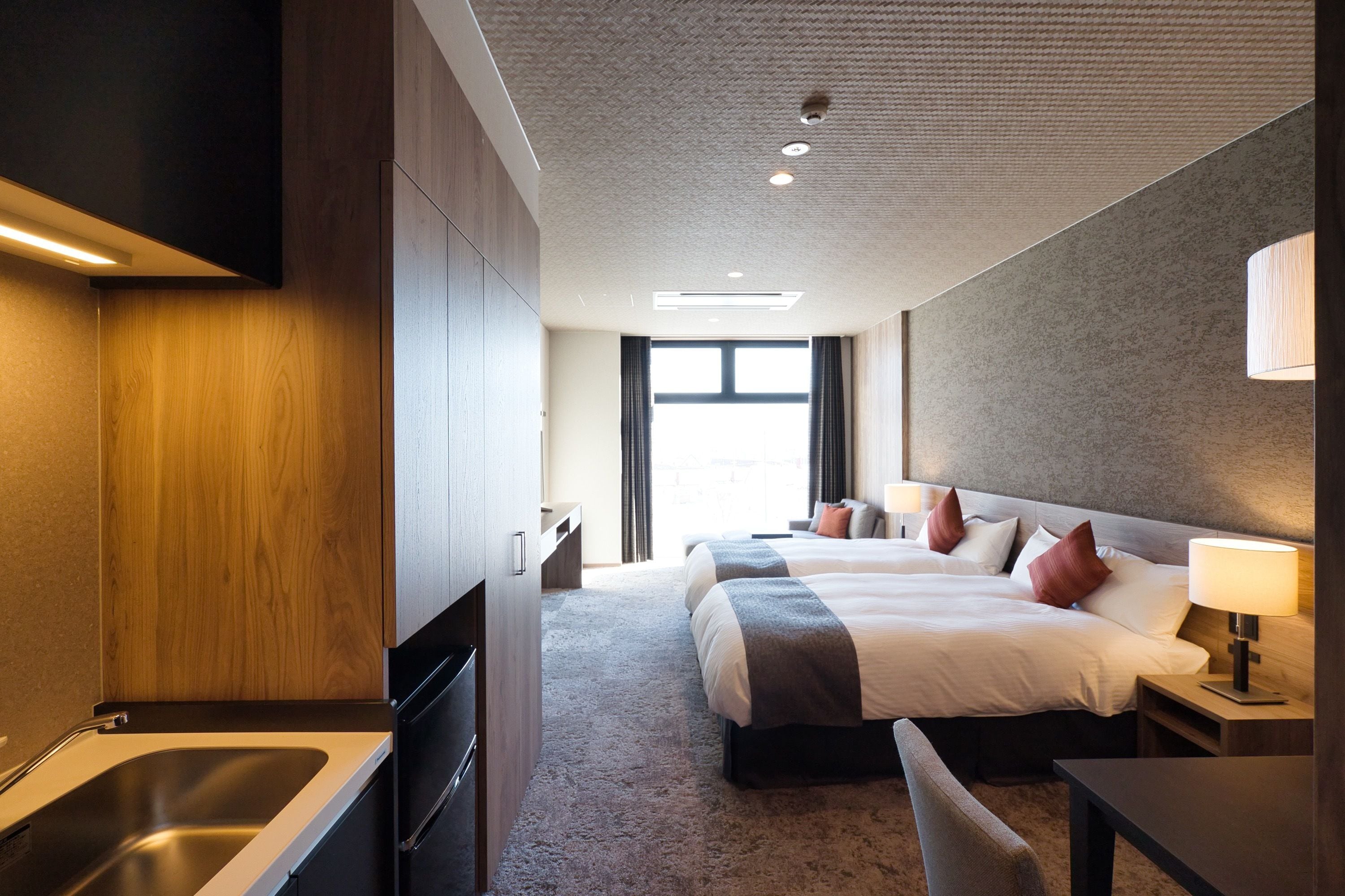 A modern hotel room with a kitchenette, two beds, a large window, and a view of the cityscape.