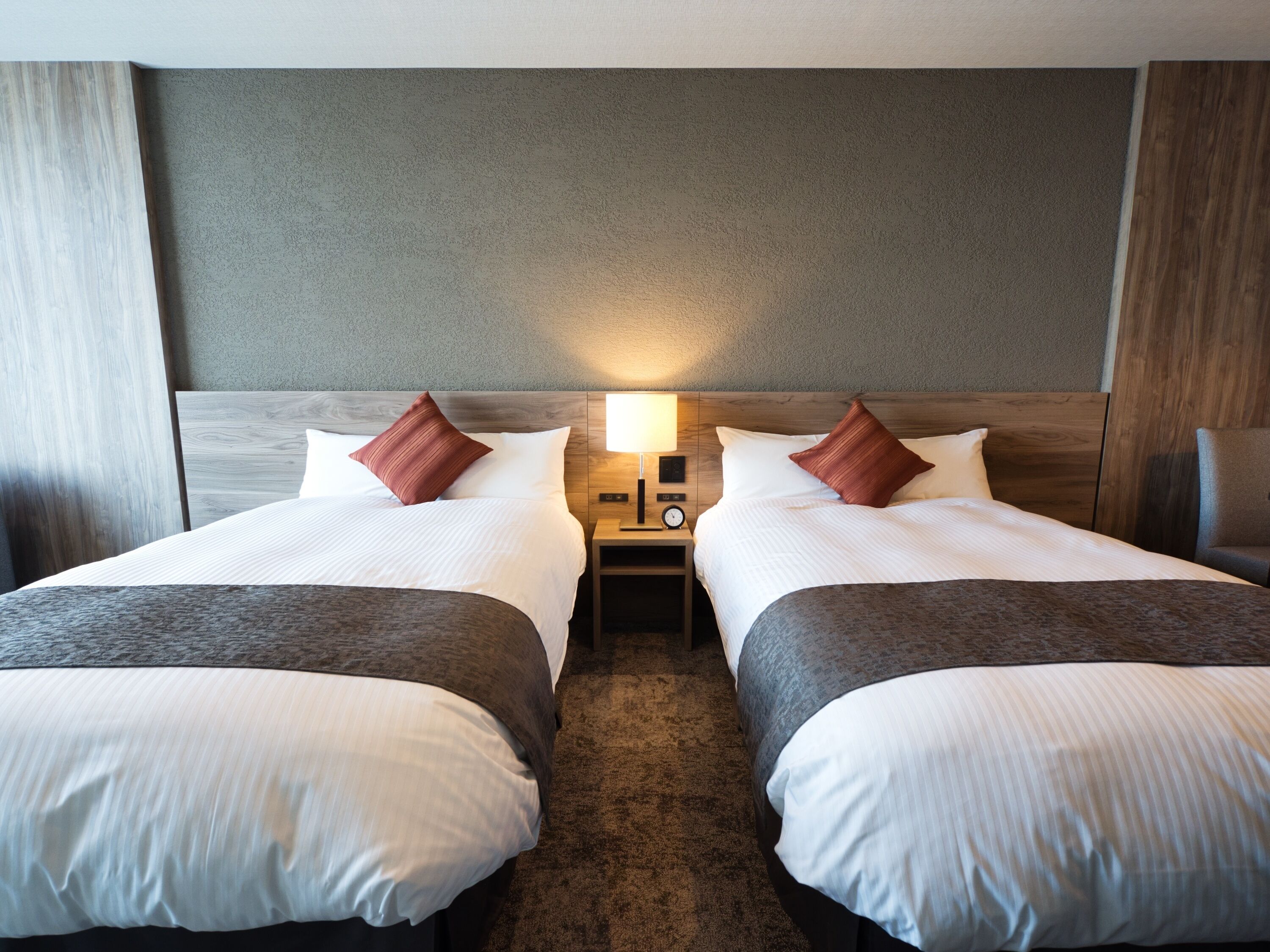 A hotel room with two beds, each with white linens and brown throw blankets. There is a nightstand between the beds with a lamp and a clock.