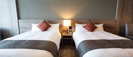 A hotel room with two beds, each with white linens and brown throw blankets. There is a nightstand between the beds with a lamp and a clock.