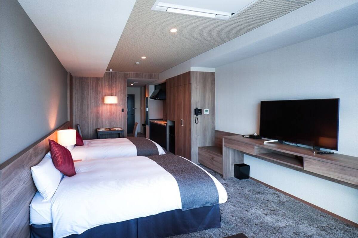 Ein Hotelzimmer mit zwei Betten, einem Fernseher, einem Schreibtisch und einer Küchenzeile.