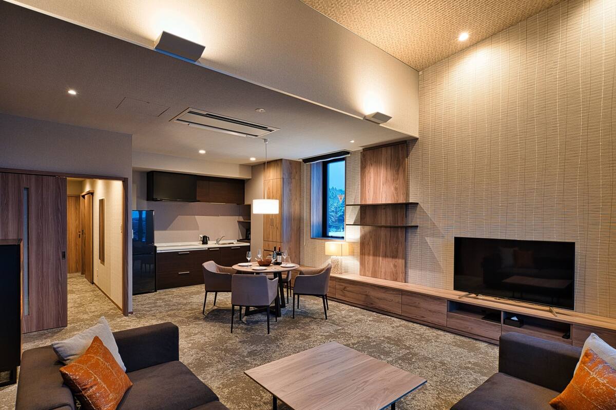 Ein modernes Wohnzimmer mit einer Sofagarnitur, einem Couchtisch und einem an der Wand befestigten Fernseher. Es gibt einen Essbereich mit Tisch und Stühlen, und im Hintergrund eine Küche.