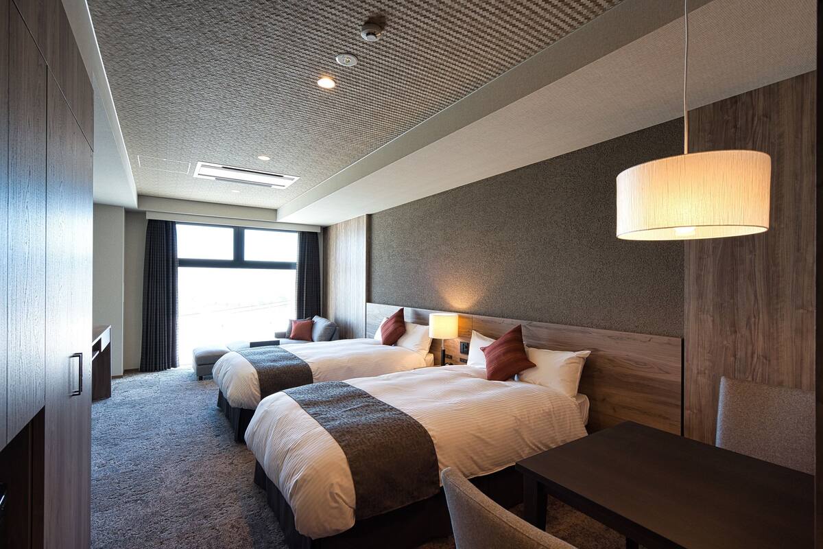 Ein Hotelzimmer mit zwei Betten, einem Schreibtisch, einer Lampe und Blick ins Freie.