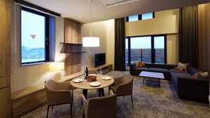 Moderni hotellihuone, jossa on ruokailutila, sohva ja suuri ikkuna, josta avautuu näkymä kaupunkiin.
