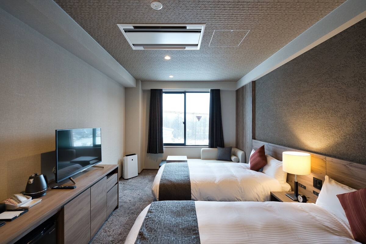 Ein Hotelzimmer mit Bett, Nachttisch, Lampe, Fernseher und einem Fenster mit Vorhängen.