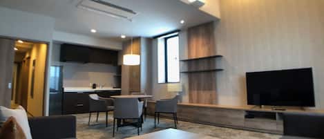 A modern living room with a sofa, coffee table, and TV. There is a dining area with chairs and a table. The room has a kitchen in the background with cabinets and a TV.