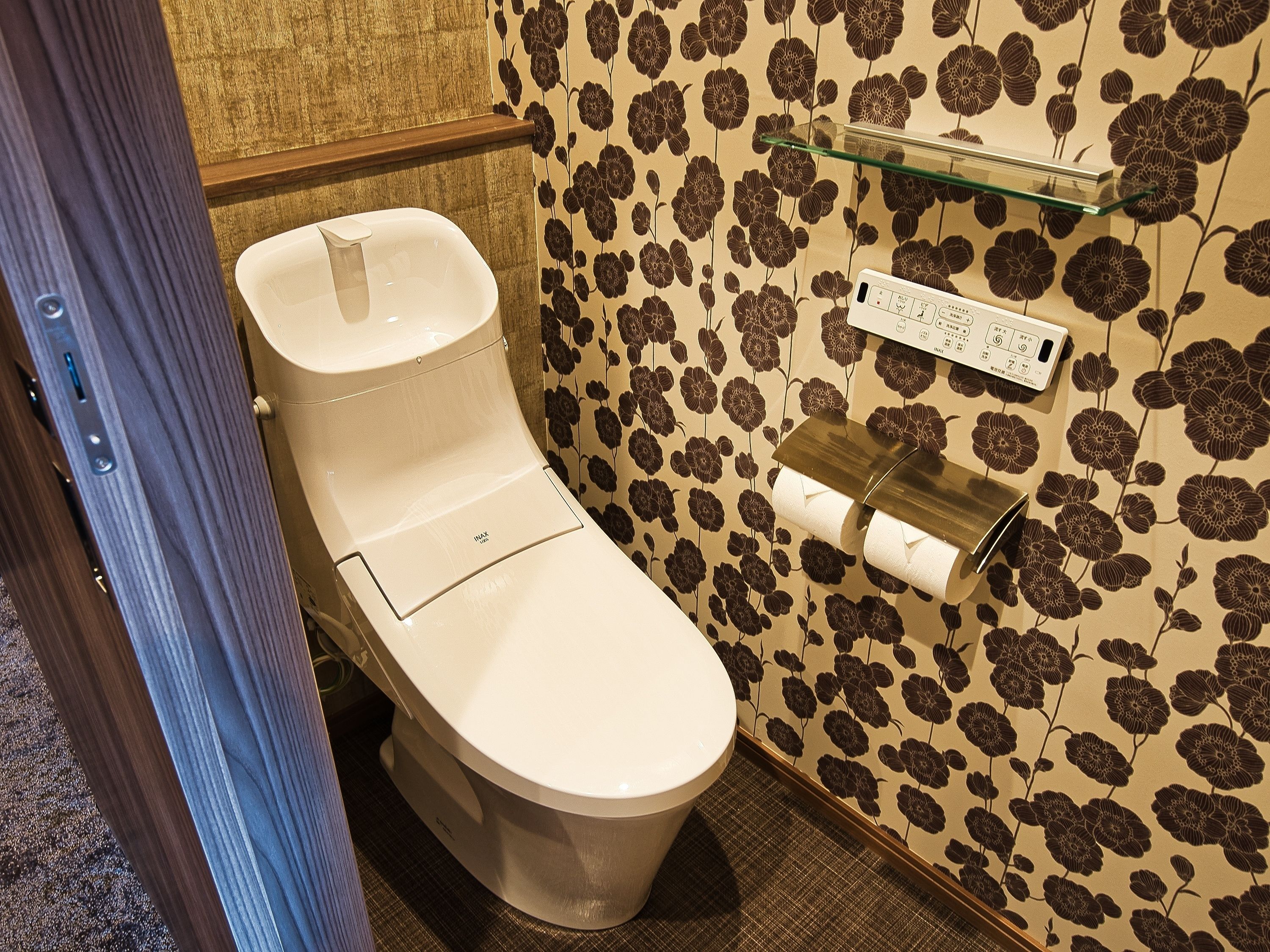 A toilet with a flush handle, a toilet paper holder, and a wall with a floral pattern.