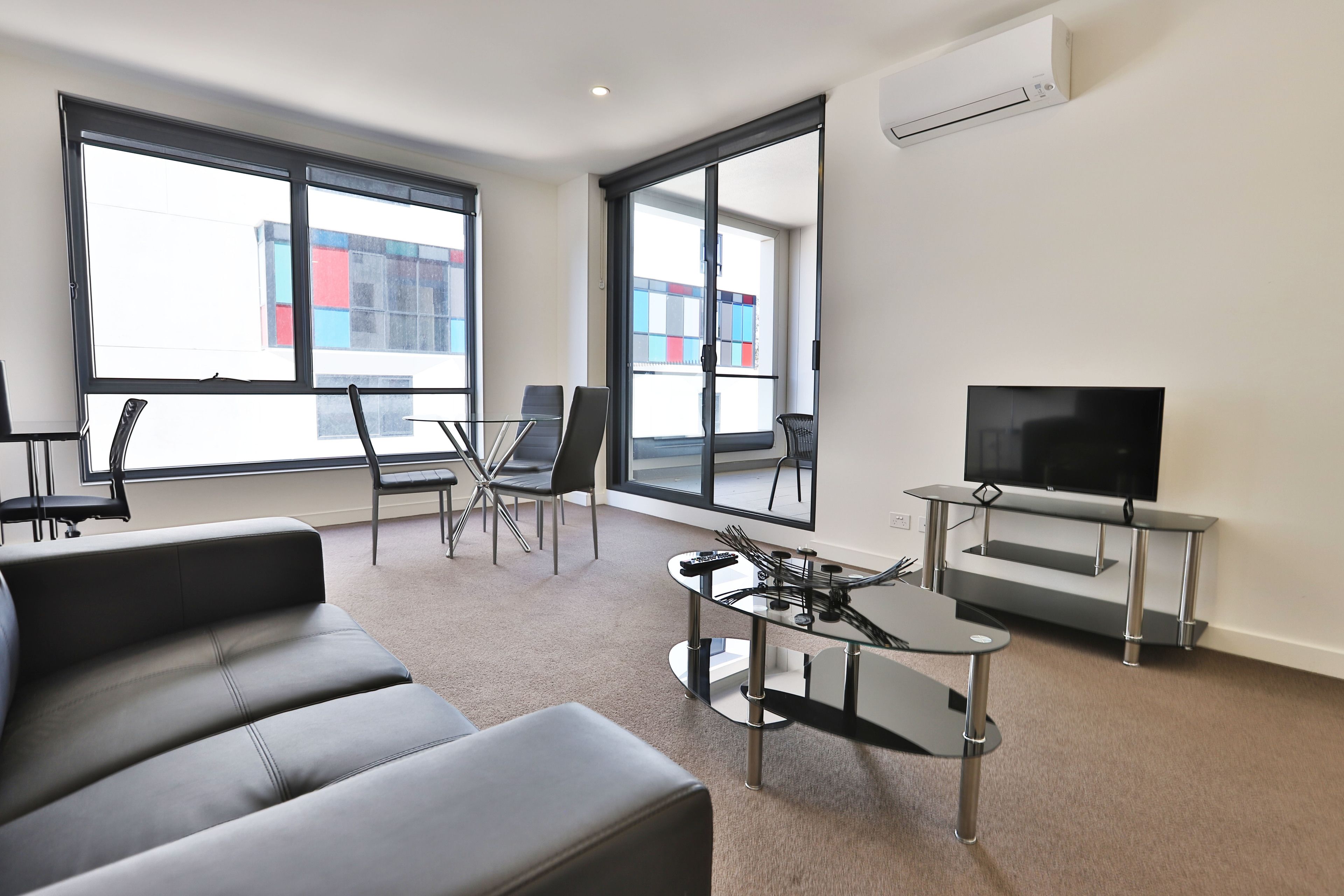 Una sala de estar moderna con un sofá, una mesa de centro de vidrio, un televisor sobre un mueble bajo y ventanas grandes que ofrecen vistas de edificios.