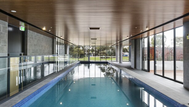 A long indoor swimming pool with clear blue water, wooden ceiling panels, and large windows along one side. - Melbourne Knox Central Apartment Hotel (Wantirna South)