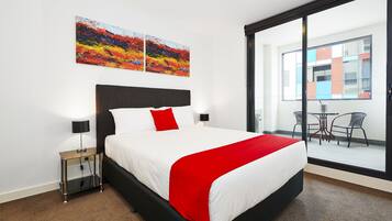 Habitación de hotel con cama, mesita de noche, una manta roja y vistas al balcón exterior.