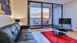A modern living room with a leather sofa, a red rug, a TV, and a balcony with a sunset view. - Melbourne Knox Central Apartment Hotel (Wantirna South)