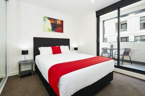 A modern hotel room with a large bed, a red throw blanket, a black headboard, a nightstand with a lamp, and a large window with a view of the outside. - Melbourne Knox Central Apartment Hotel (Wantirna South)