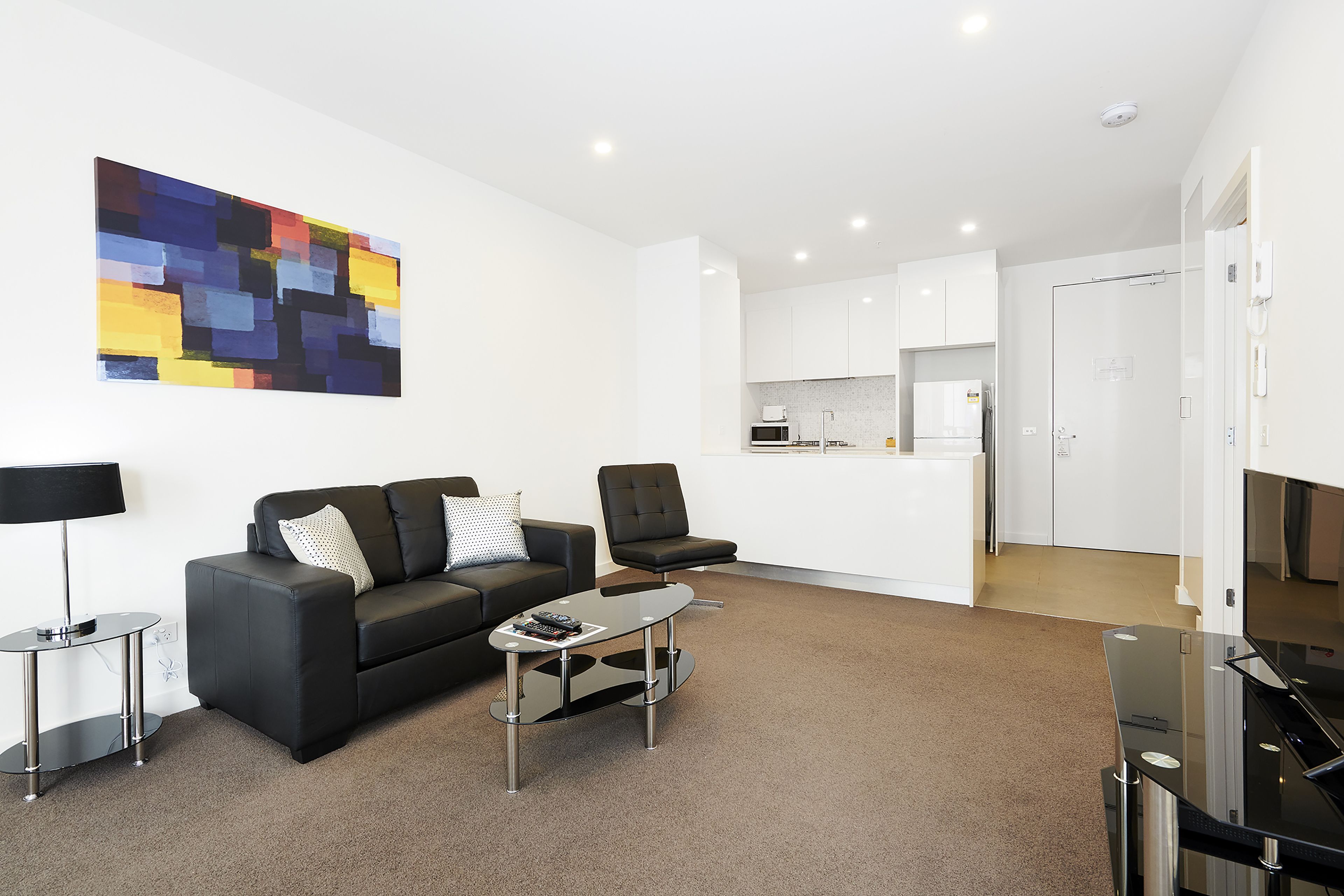 A modern living room with a black sofa, a glass coffee table, and a black armchair. There is a kitchen area visible through an open door.
