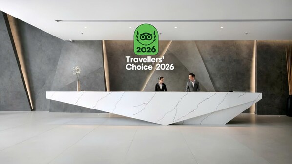A modern reception desk with a marble design, two individuals standing behind it, and a logo for 'Travelers' Choice 2026' on the wall. - M Hotel Saigon (Ho Chi Minh City)