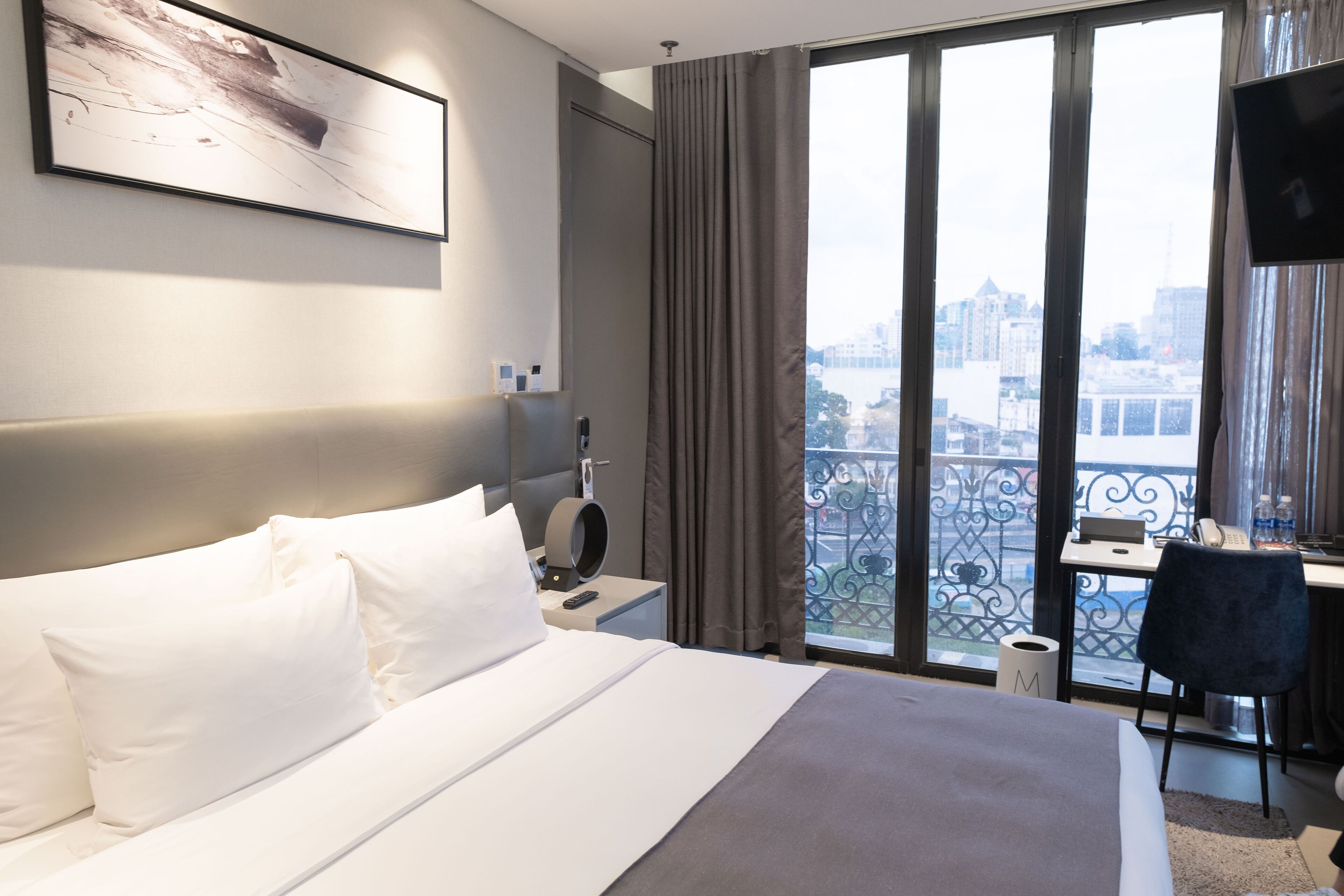 A hotel room with a large bed, a desk with a chair, a TV, and a view of the city through large windows.
