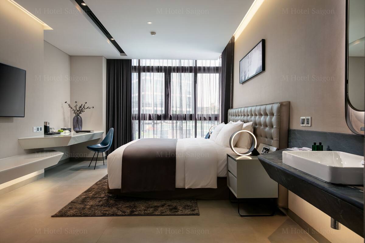 A modern hotel room with a large bed, bedside table with a mirror, a vanity with a sink, and a desk with a chair.