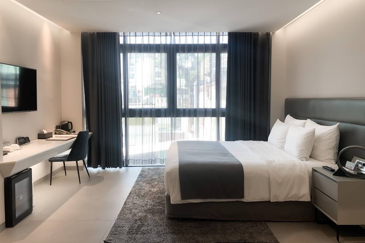 A modern hotel room with a large bed, a desk, a television, and a view of the city through large windows.