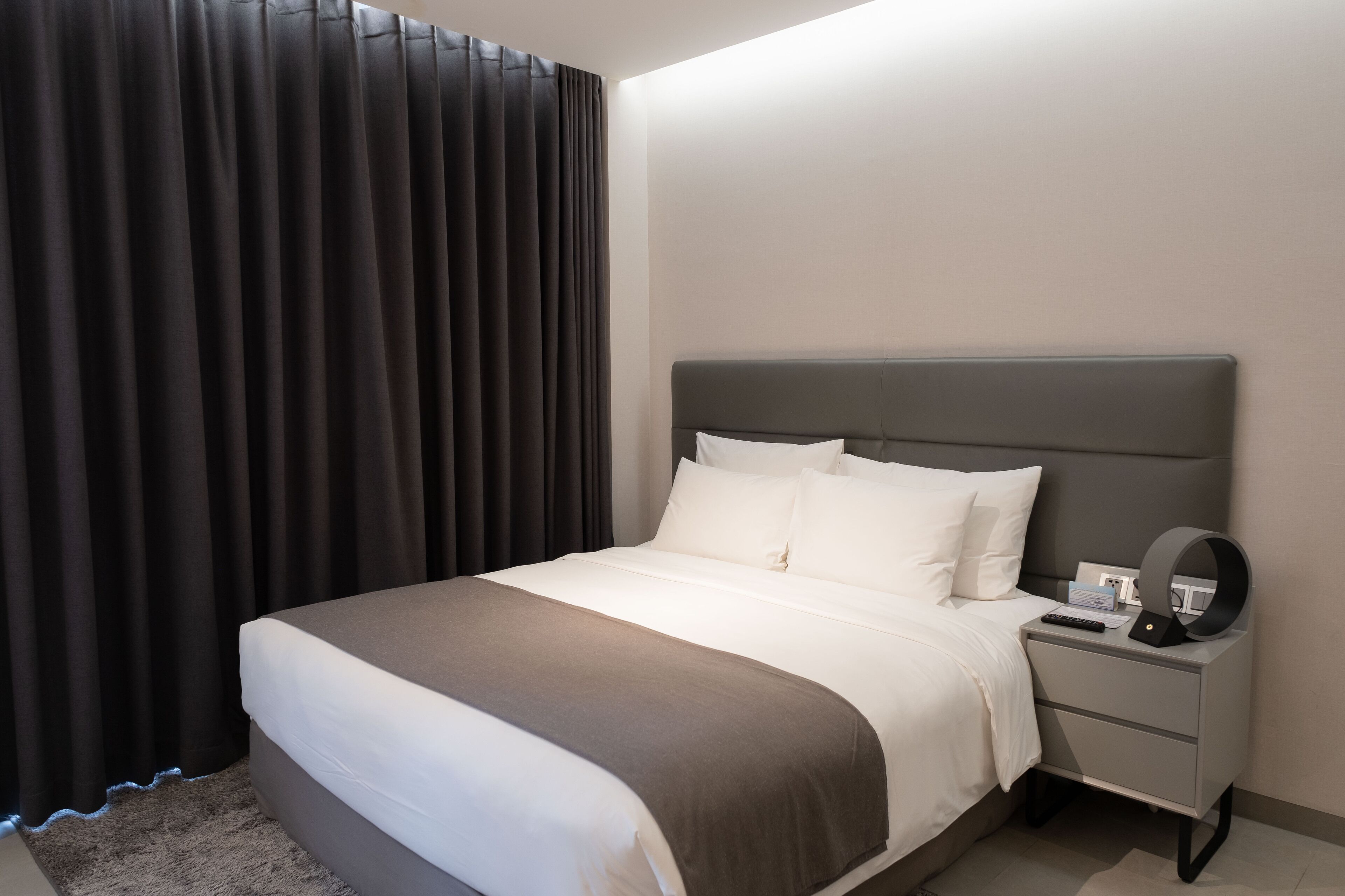 Ein modernes Hotelzimmer mit einem großen Bett, einem Nachttisch mit Lampe und Blick ins Freie durch transparente Vorhänge.