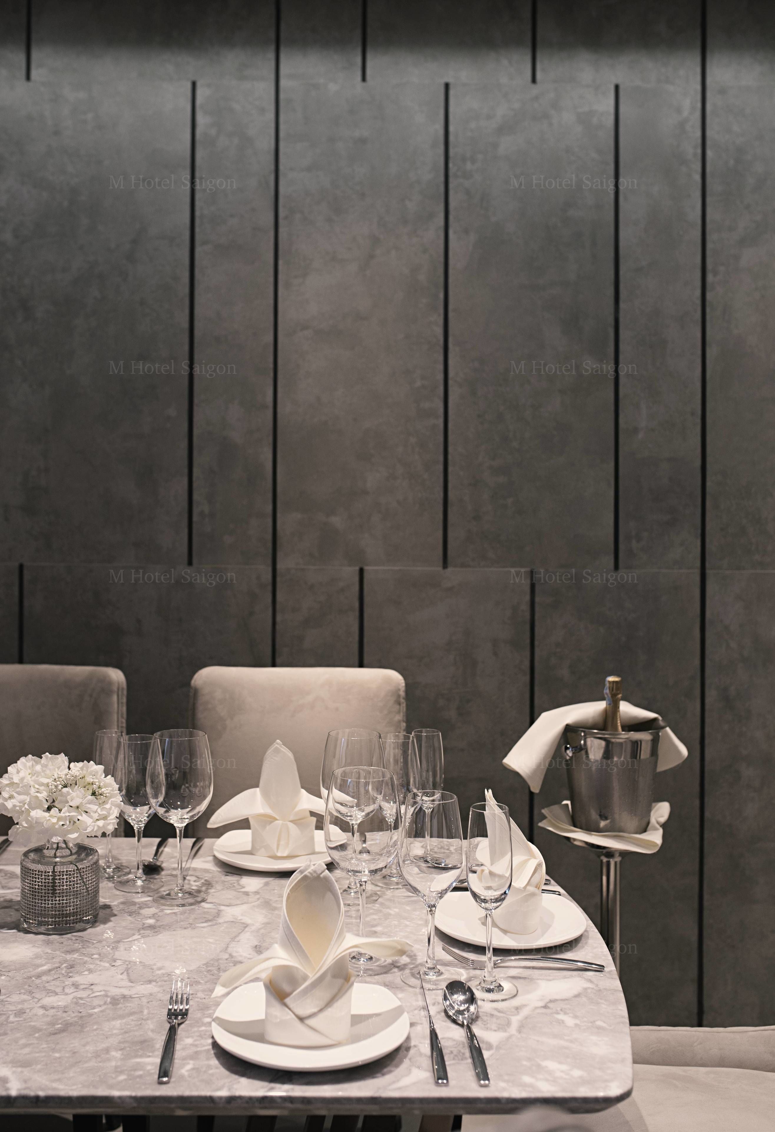 Una mesa de comedor dispuesta con platos blancos, servilletas y cubiertos, con un jarrón de flores blancas y una botella de champán a un lado.