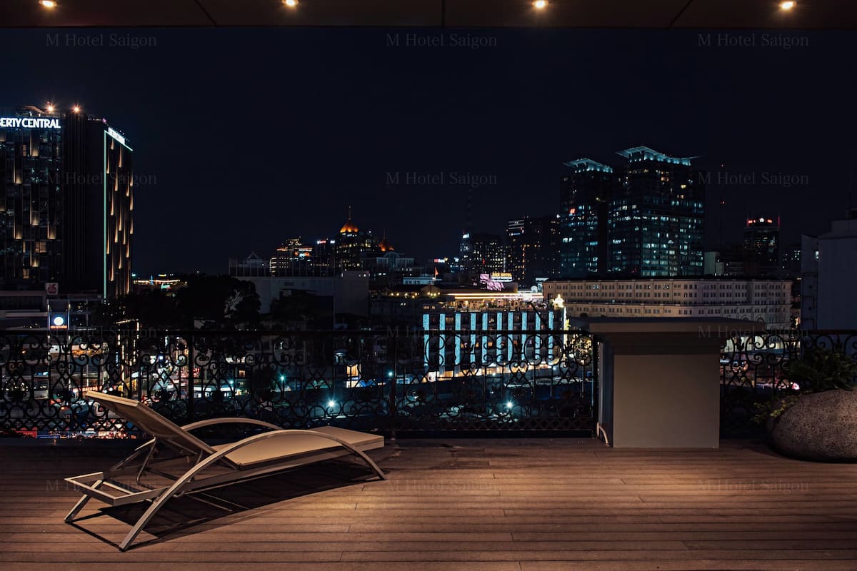 A rooftop terrace with a lounge chair, a small seating area, and a view of the city skyline at night.