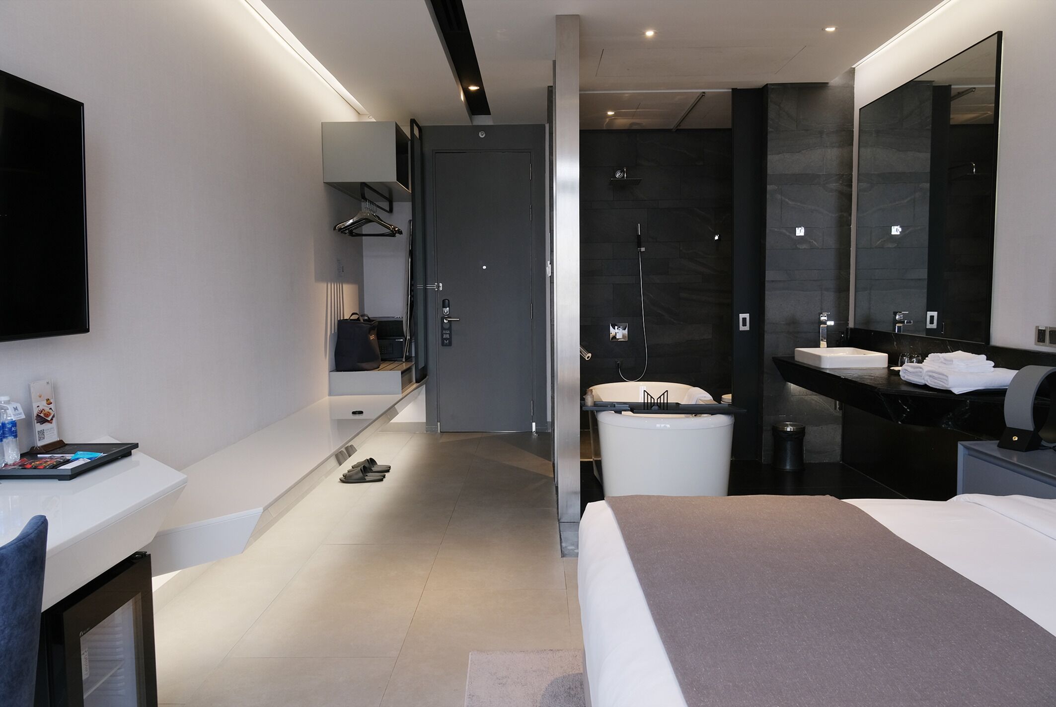 A modern hotel room with a large bed, a bathroom with a shower, a vanity with a mirror, and a desk with a chair.