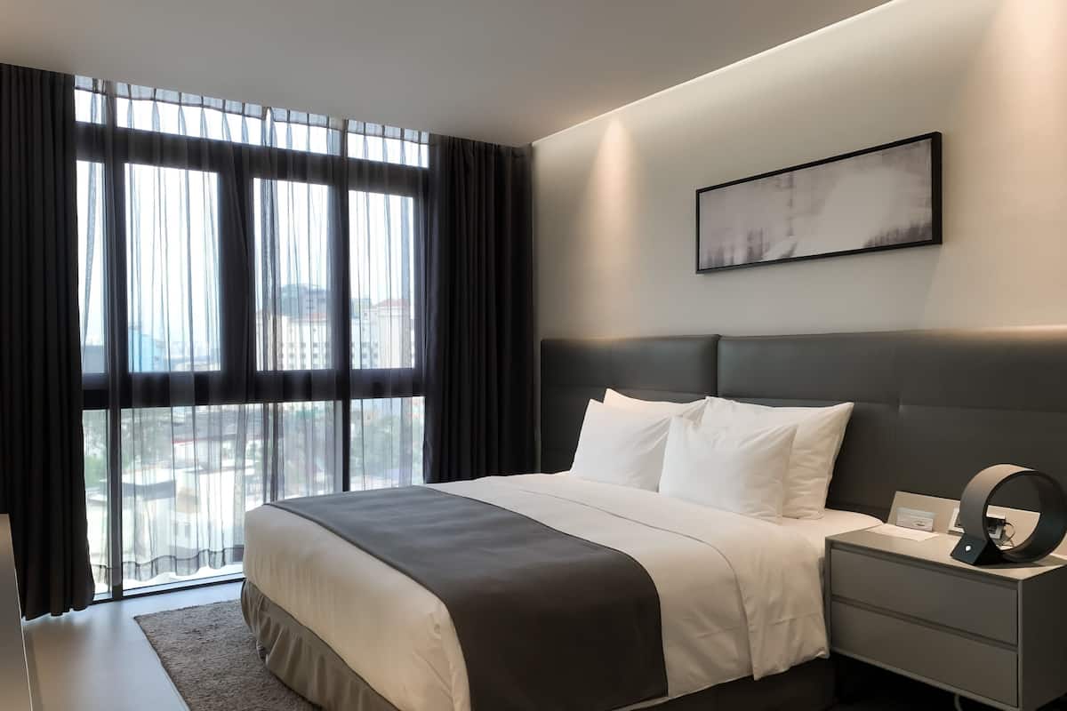 A modern hotel room with a large bed, a bedside table with a lamp, and a view of the city through floor-to-ceiling windows.