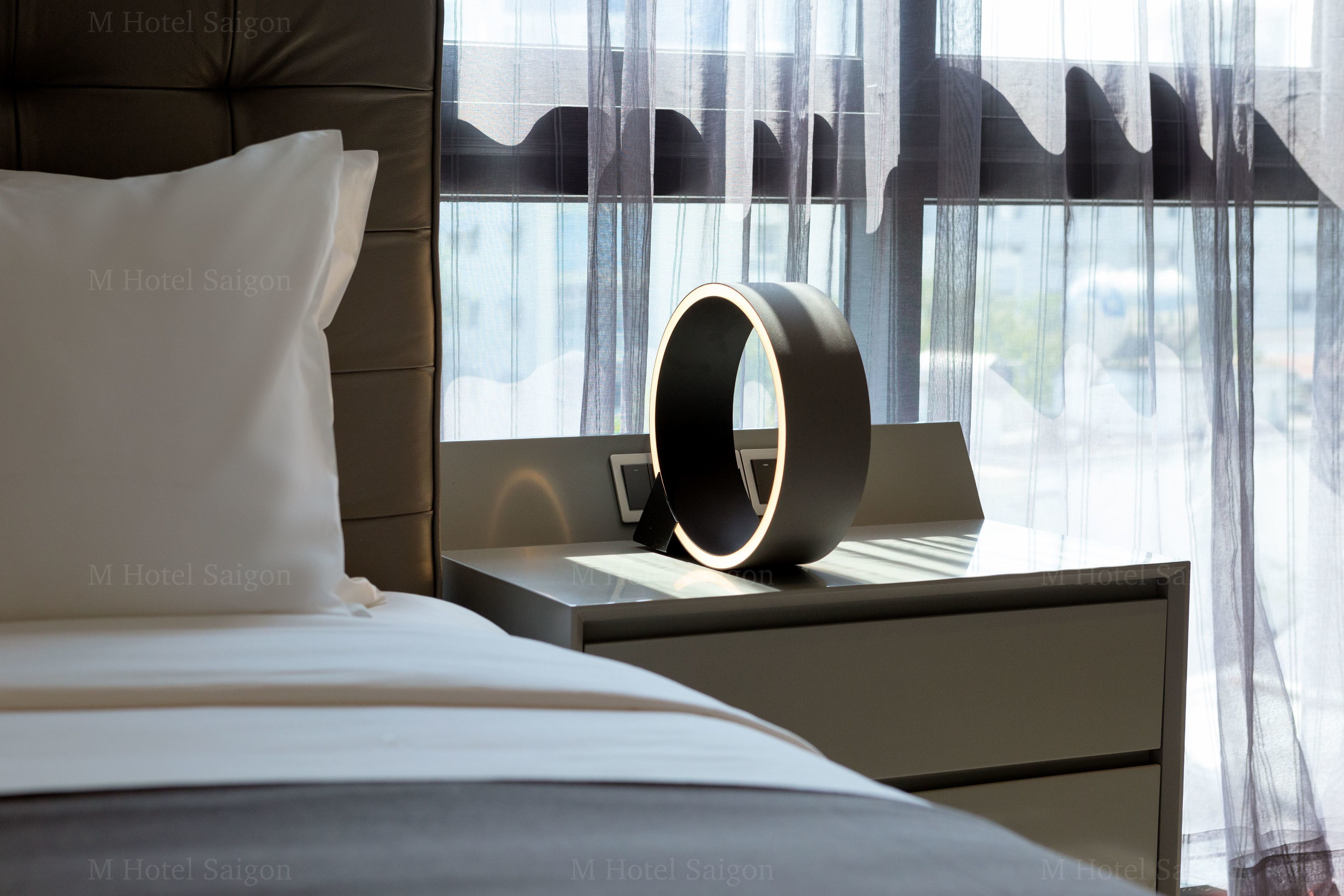 Une chambre d’hôtel avec un lit, une table de chevet dotée d’une lampe moderne et une vue dégagée à travers des rideaux légers.
