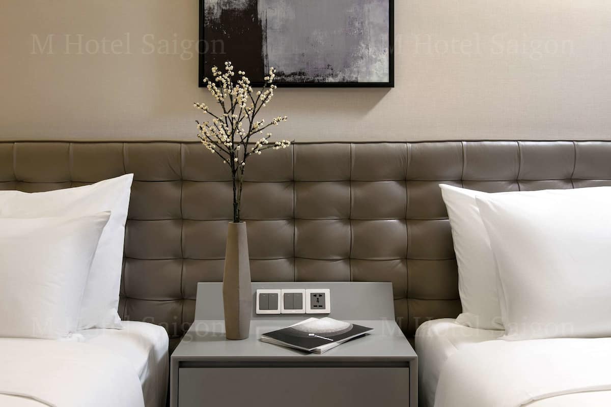 A hotel room with a tufted headboard, two beds with white bedding, a bedside table with a vase of flowers, a framed artwork on the wall, and a modern design aesthetic.