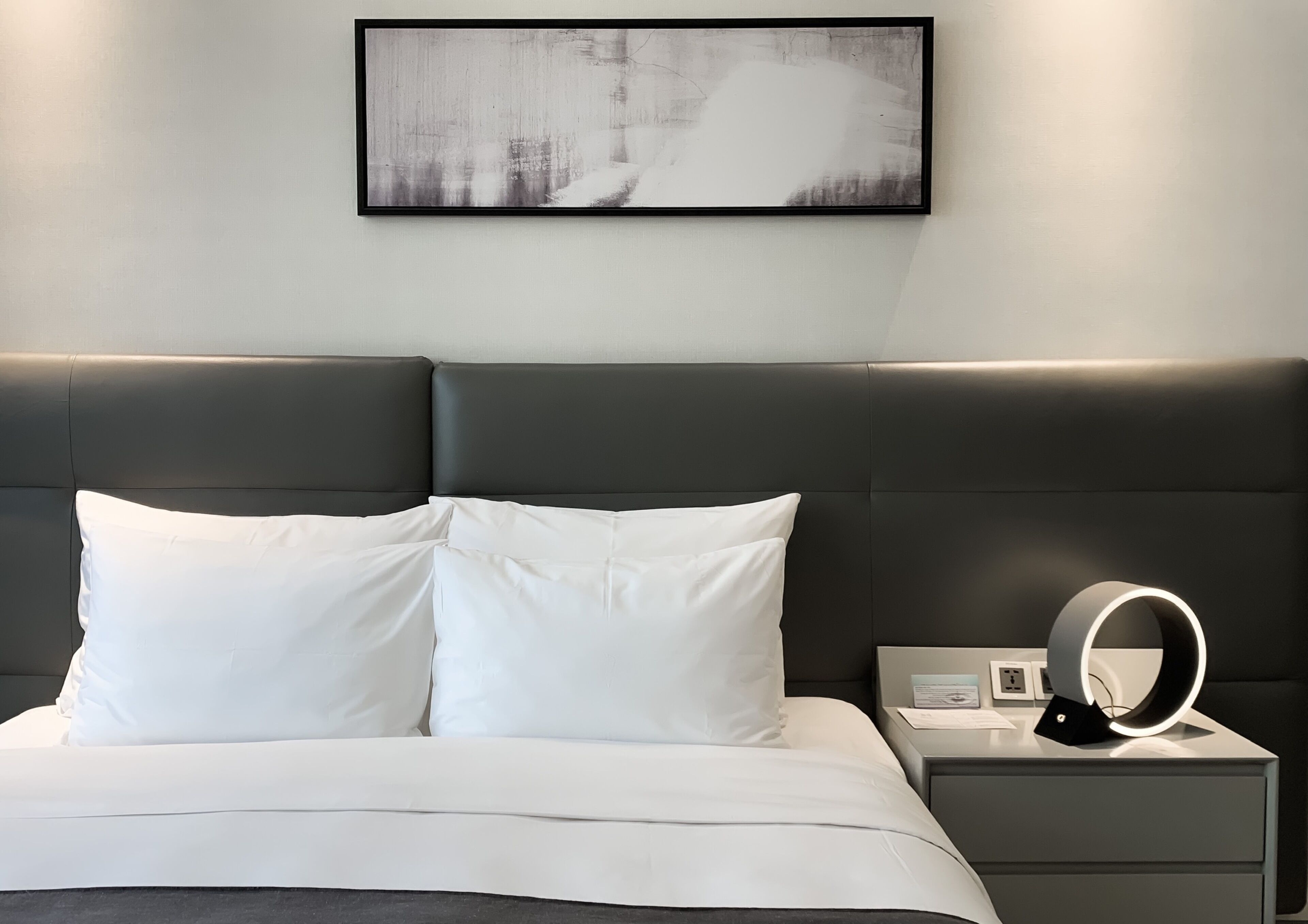 A modern hotel room with a large bed, white bedding, a dark headboard, a bedside table with a ring light, and a framed abstract artwork on the wall.