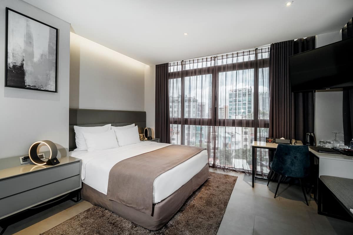 A modern hotel room with a large bed, a desk, and a view of the cityscape through sheer curtains.