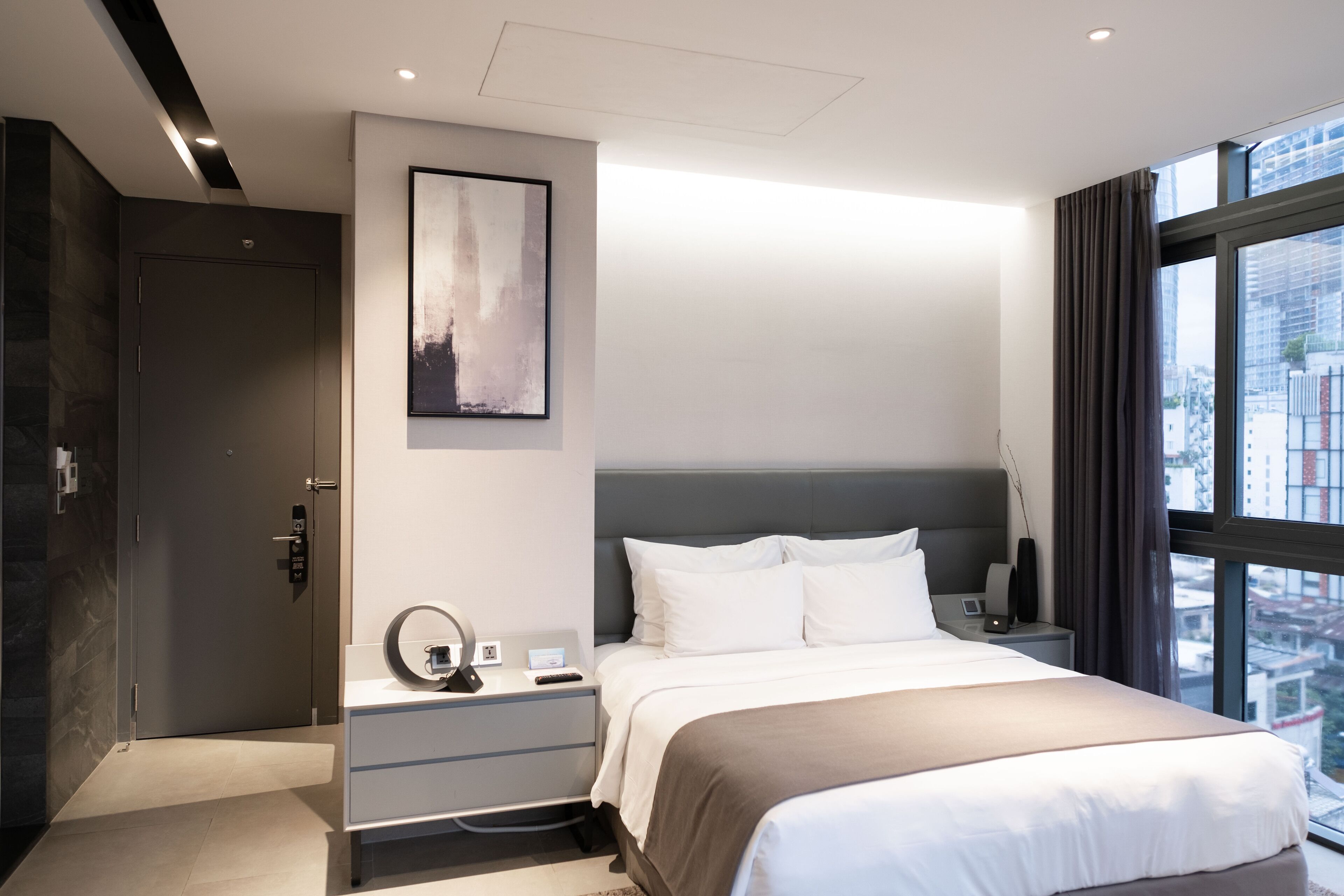 A modern hotel room with a large bed, a bedside table with a lamp, a wall-mounted artwork, and a view of the city through a large window.