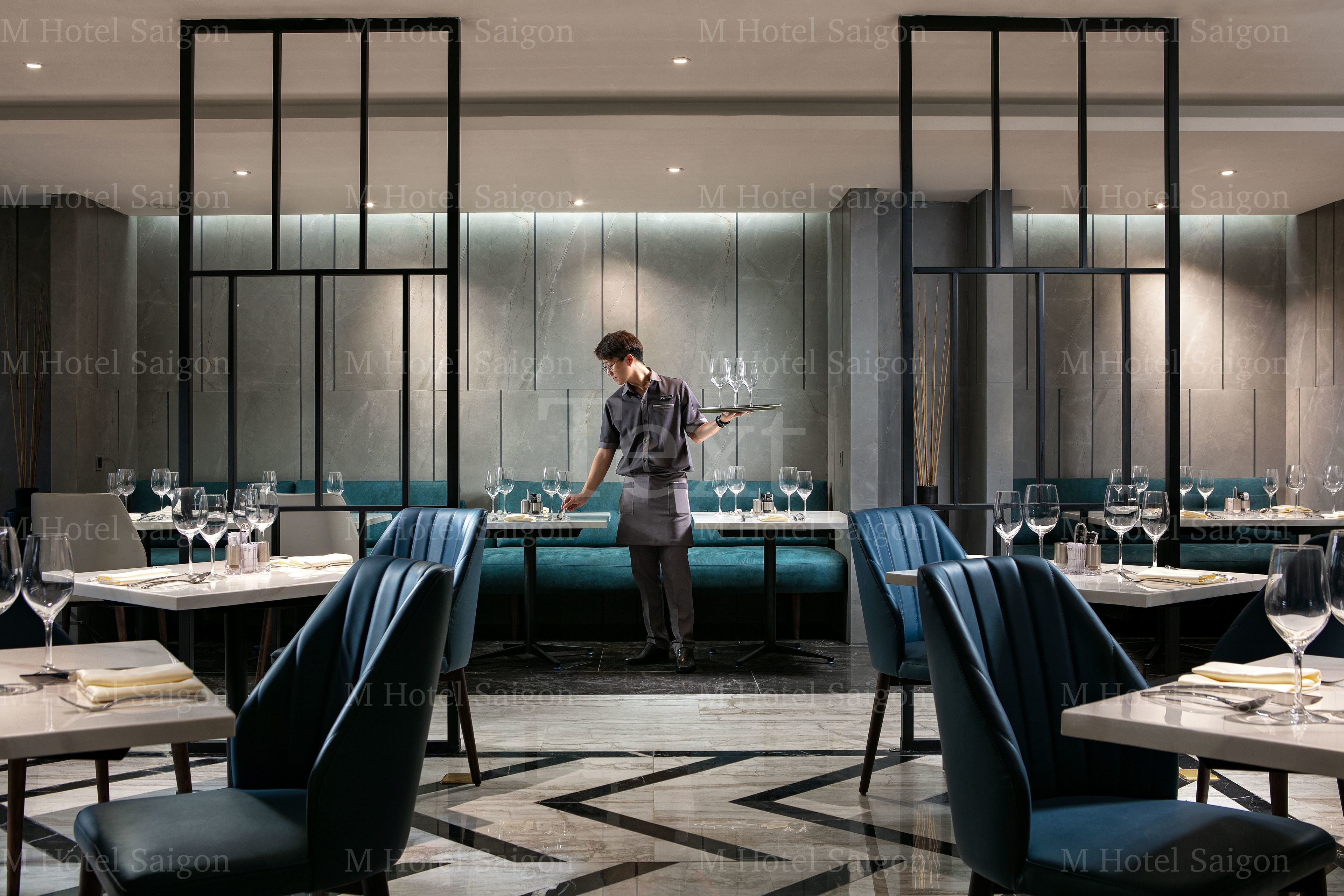 En moderne restaurant med en tjener, der bærer en bakke med glas. Bordene er dækket med vinglas, vandglas og bestik. Rummet har et moderne design med mørkeblå stole og et geometrisk gulvmønster.