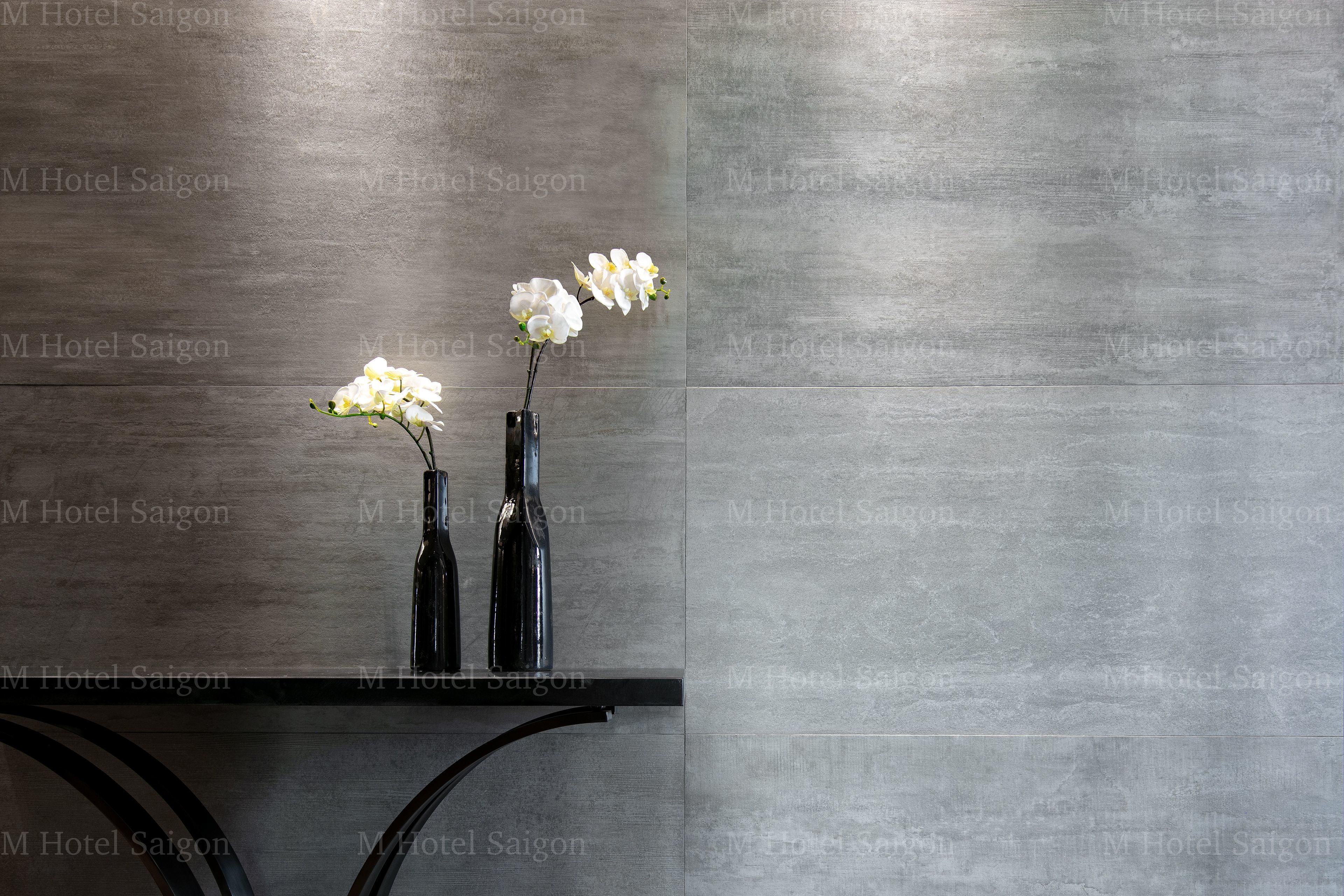 A modern interior with a grey tiled wall and two black vases with white orchids on a black shelf.