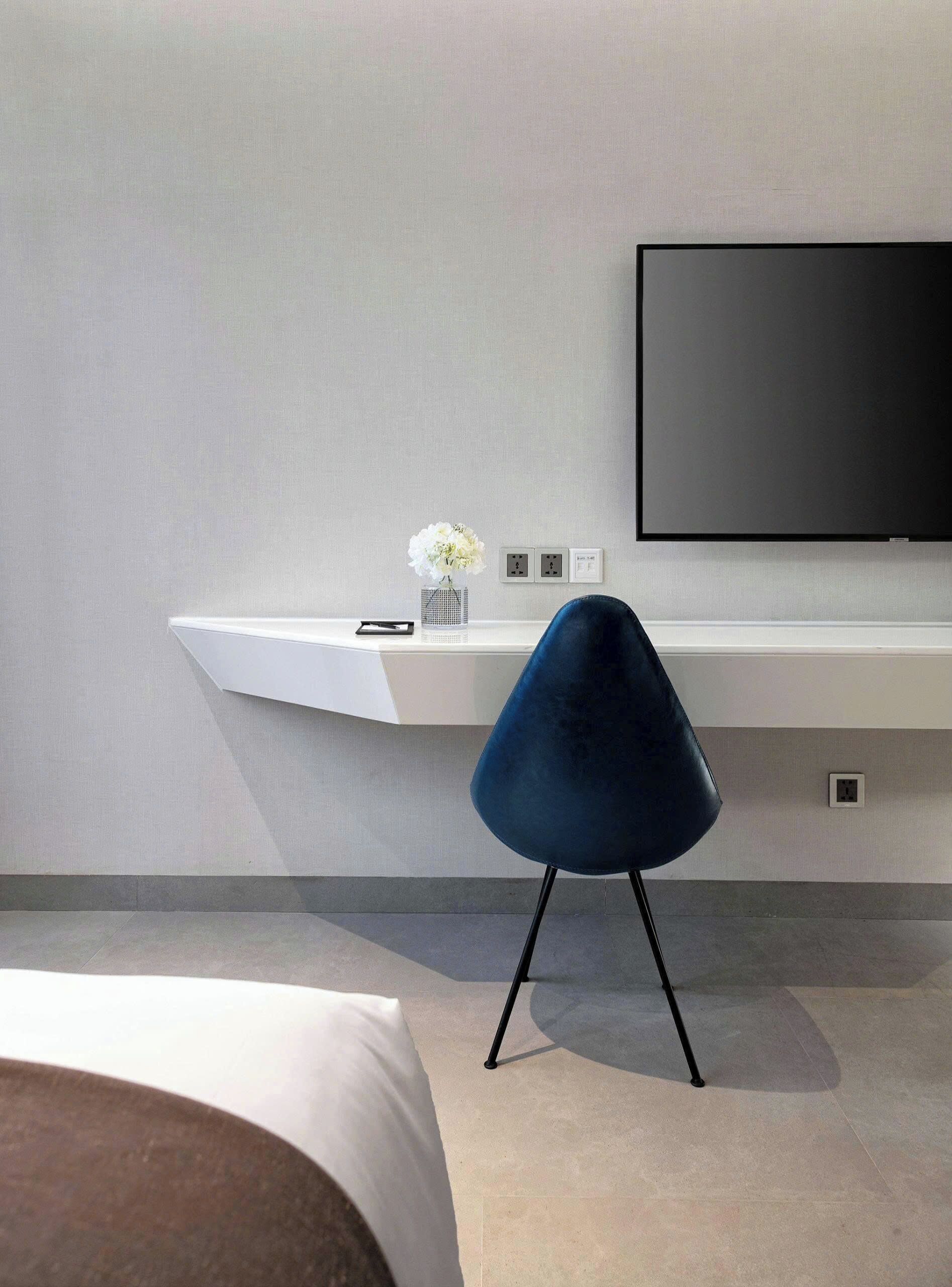 A modern hotel room with a white desk, a blue chair, a flat-screen TV, and a vase with flowers.