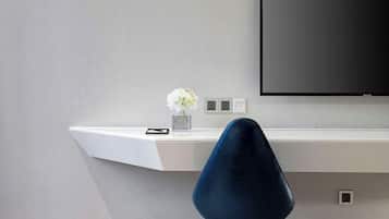 A modern hotel room with a white desk, a blue chair, a flat-screen TV, and a vase with flowers.