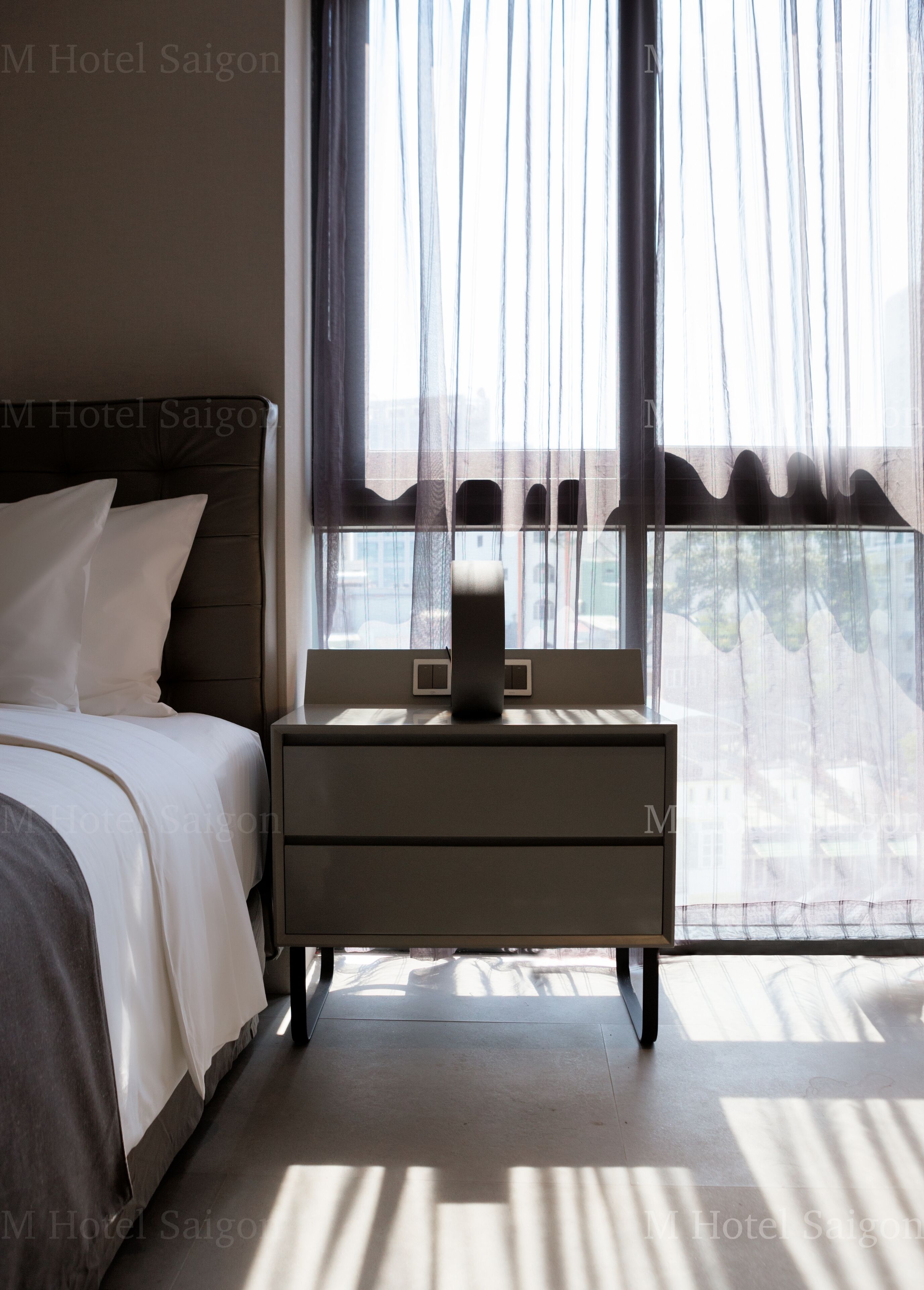 A hotel room with a bed, bedside table, and a window with sheer curtains.
