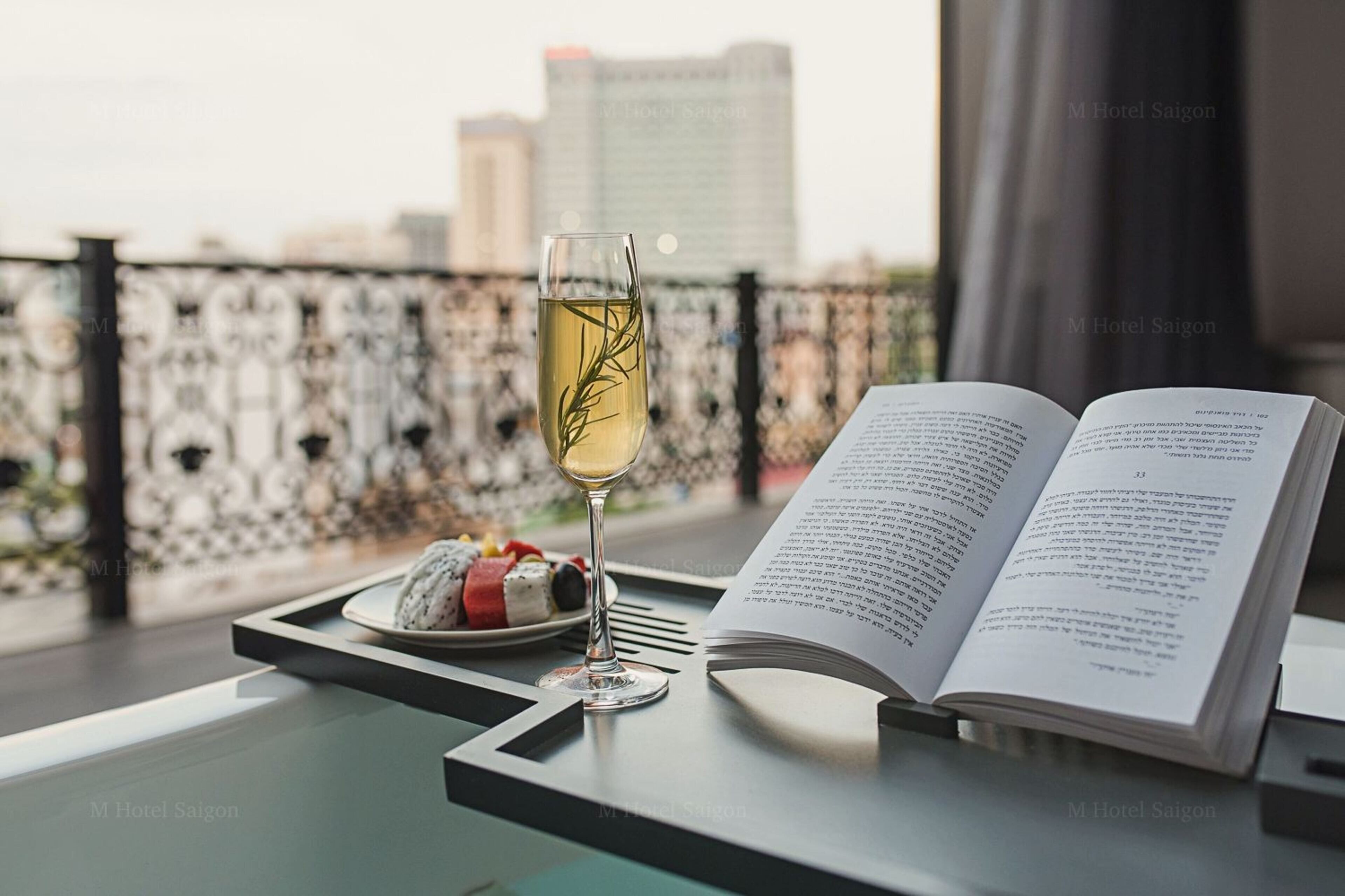 Une table avec un livre, un verre de vin et une assiette de nourriture, le tout sur un balcon offrant une vue sur la ville.