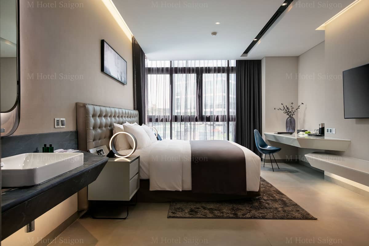 A modern hotel room with a large bed, a bedside table with a mirror, a desk with a chair, a large window with curtains, and a TV mounted on the wall.