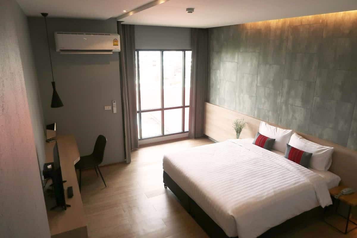 A modern hotel room with a large bed, a desk, and a chair. There is a window providing natural light and a view of the outside.