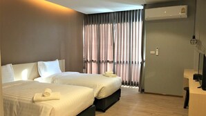 A hotel room with two beds, a desk, an air conditioning unit, and a window with curtains. - Sans Hotel (Ratchaburi)
