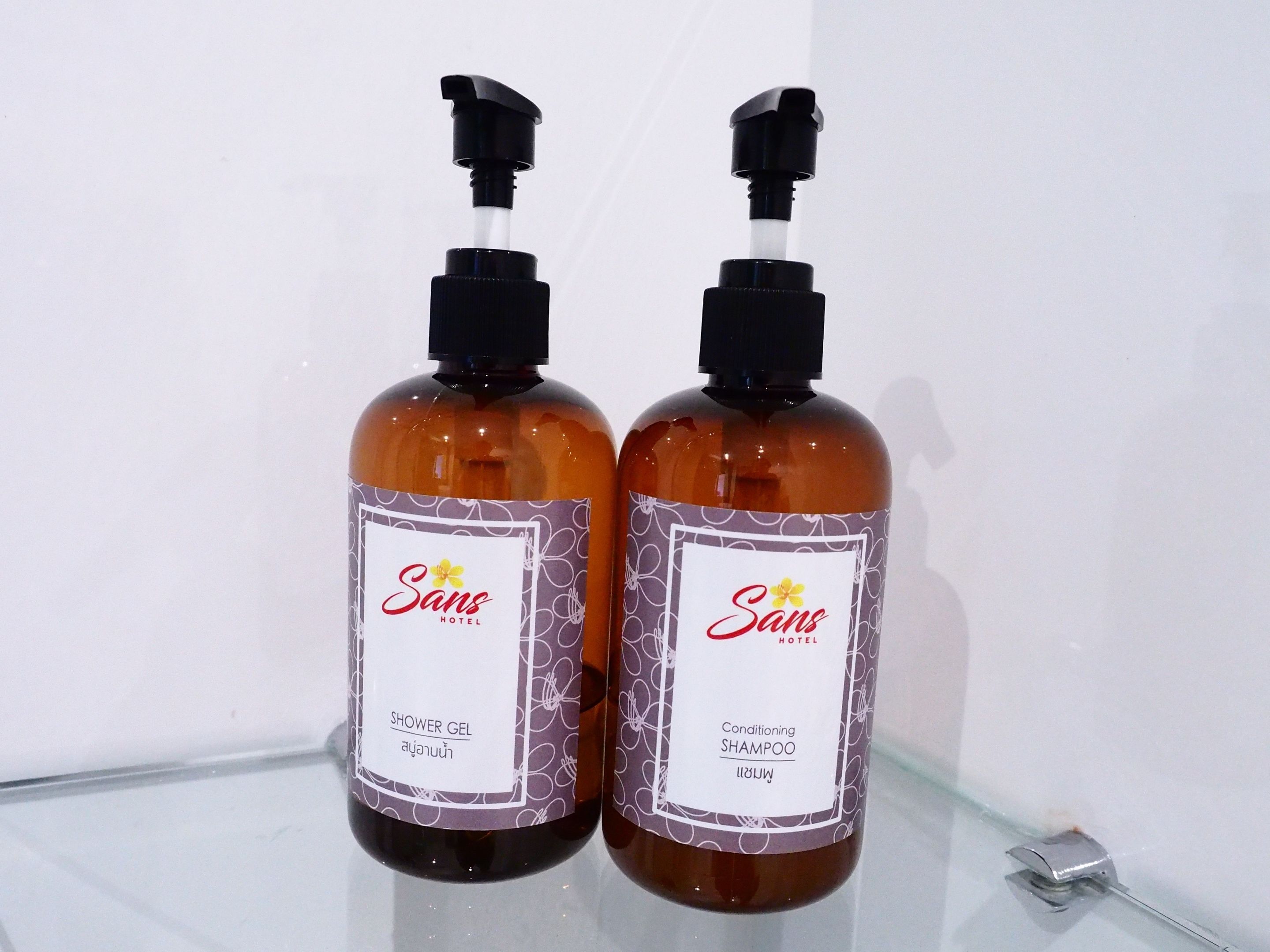 Two bottles of Sans Hotel shower gel and conditioning shampoo on a glass shelf.
