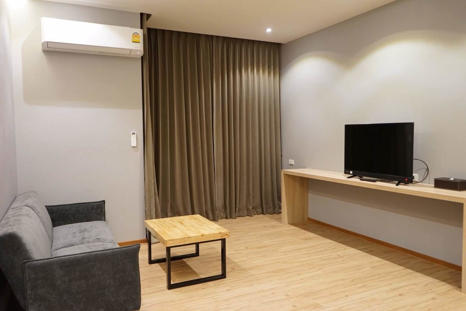 A modern living room with a grey sofa, a wooden coffee table, a TV on a wooden stand, and a large window with curtains.