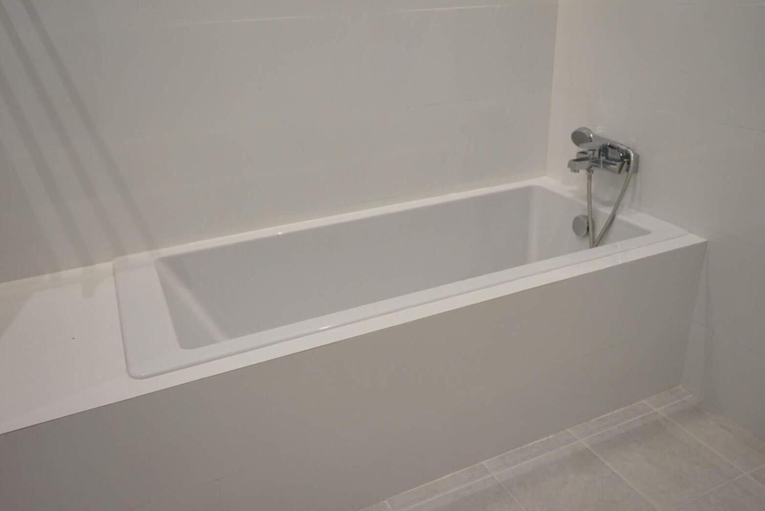 Una vasca da bagno rettangolare standard con rubinetteria cromata e soffione, posta di fronte a una parete liscia e pavimento piastrellato.