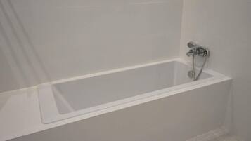 Una vasca da bagno rettangolare standard con rubinetteria cromata e soffione, posta di fronte a una parete liscia e pavimento piastrellato.