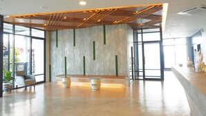 A spacious lobby with a modern design, featuring a concrete wall with green accents, a wooden ceiling with a geometric pattern, and a reception desk with two decorative vases. - Sans Hotel (Ratchaburi)
