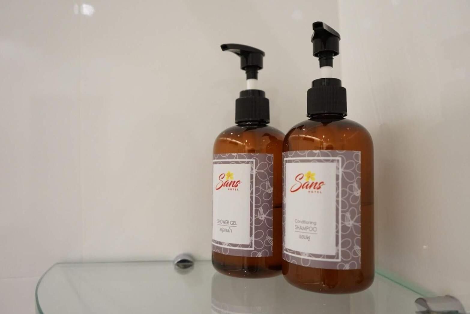 Dois frascos de gel de banho e shampoo condicionador Sans hotel sobre uma prateleira de vidro.