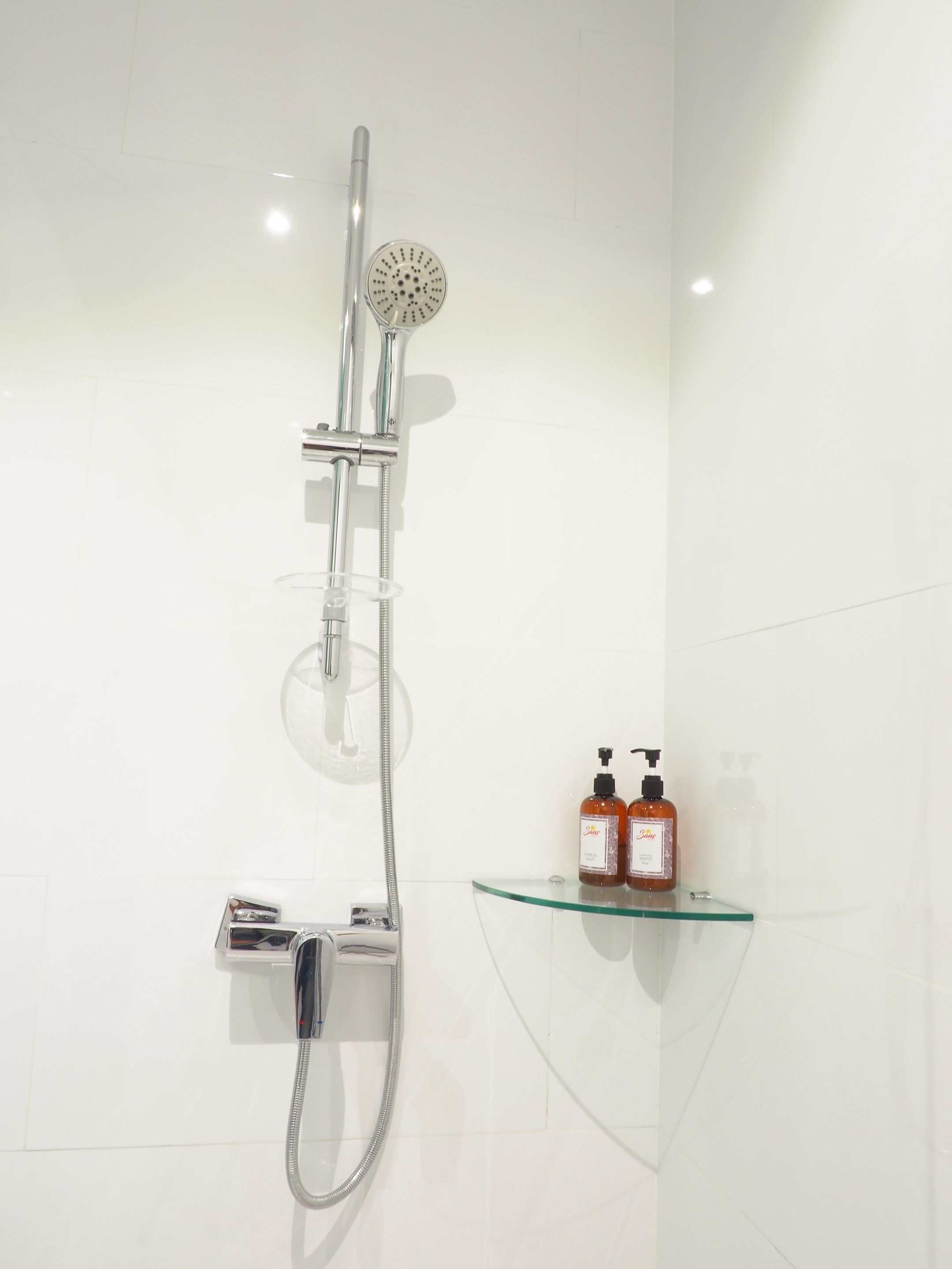A modern shower with a handheld showerhead and a fixed rain showerhead, mounted on a wall with a glass shelf holding two bottles of shampoo.